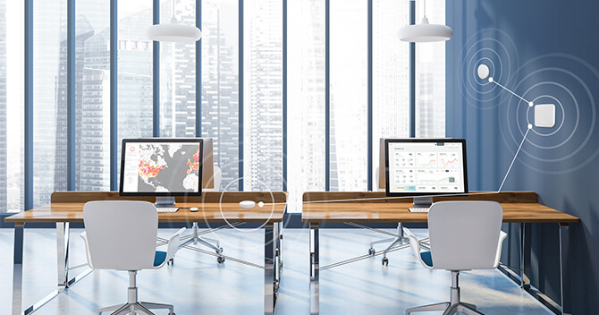 Two office desks with screens showing the Airthings Dashboard and radonmap.com with the Airthings Wave and Airthings Hub on the wall