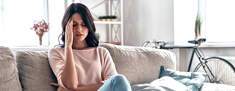 Woman sitting on couch at home with hand to her head indicating a headache